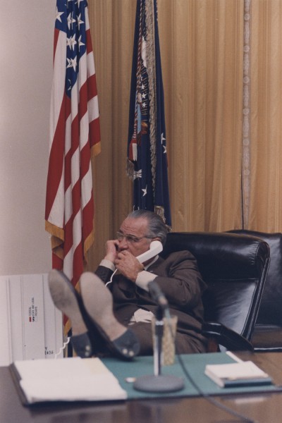 Photographic print of LBJ seated in a leather chair, holding a telephone to his ear with feet propped on a large desk, American and presidential flags behind him