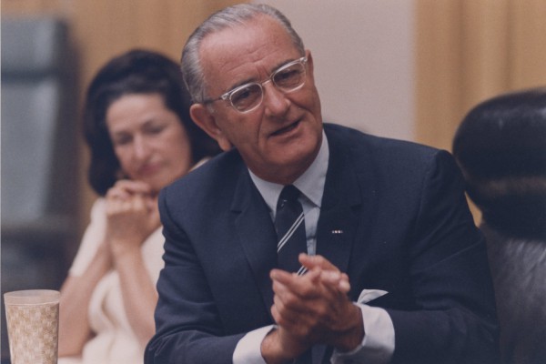 Photographic print of LBJ in a navy suit and glasses, hands clasped, speaking at a table. Lady Bird sits blurred in the background, hands folded