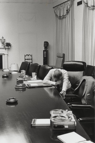 Photographic print of LBJ seated alone at a long conference table, head resting in his hands, with a reel-to-reel recorder and papers nearby