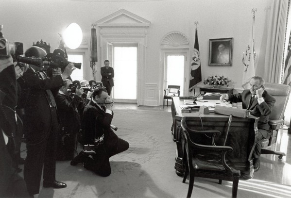 Photographic print of LBJ speaking on the phone at the Oval Office desk while a crowd of press photographers and camera operators record him