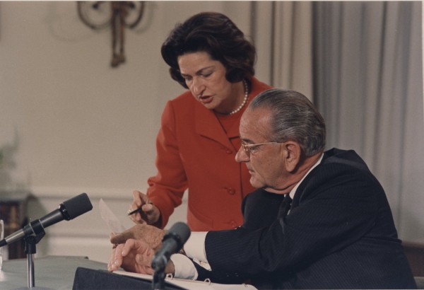 Photographic print of Lady Bird Johnson, in a red jacket and pearls, holding a pen over speech papers at a podium where a suited man sits reviewing documents