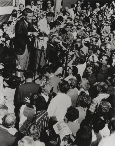 Photographic print of a man speaking at a microphone-lined podium above a dense outdoor crowd, with a "Bye-Bye" campaign sign visible in the background