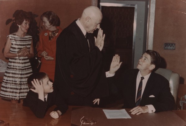 Photographic print of a man seated at a desk raising his right hand before a robed official, with a young boy and two women looking on in an indoor setting