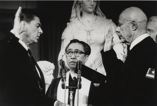 Photographic print of Ronald Reagan raising his right hand during a swearing-in ceremony, facing an official with raised hand at microphones, with a robed figure standing between them
