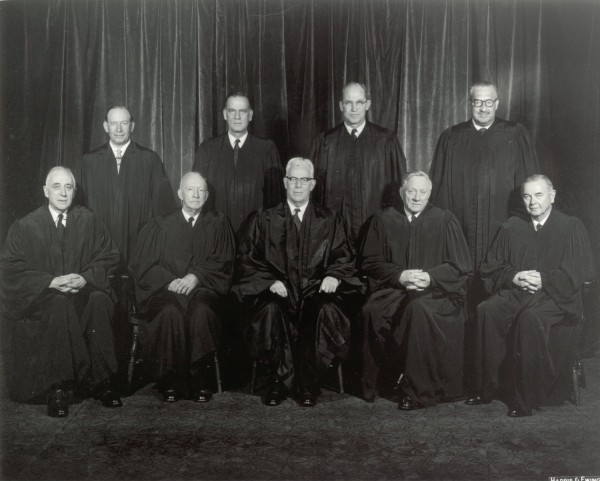 Photographic print of nine U.S. Supreme Court justices in black robes, five seated in front and four standing behind, posed before a draped curtain backdrop