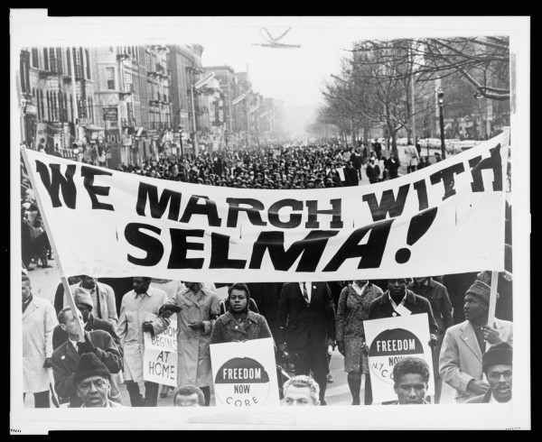 Photographic print of a large crowd filling a city street behind a banner reading "We March With Selma!"; marchers carry signs reading "Freedom Now" and "CORE"