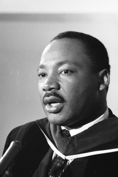 Black-and-white photograph of a man in academic regalia speaking at a microphone, shown in close-up from the chest up