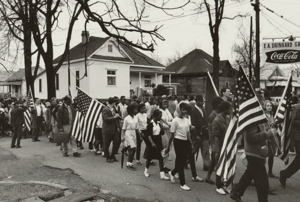 Black-and-white photograph of a large group of marchers on a residential street, several carrying American flags, with a Coca-Cola sign visible at right