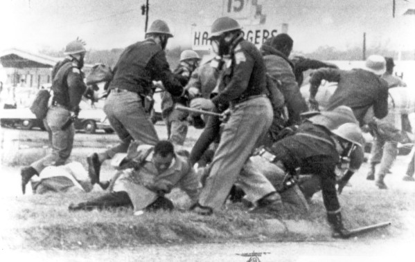 Black-and-white photograph. Helmeted officers in uniform strike and surround civilians who have fallen to the ground, with a commercial street visible in the background