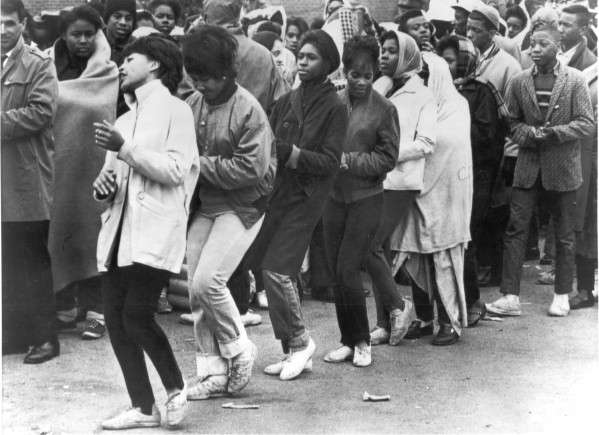 Black-and-white photograph of a large crowd of young people standing close together outdoors, dressed in coats and jackets, some with arms linked