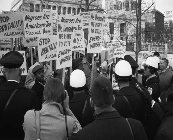 Black-and-white photograph of a crowd of demonstrators holding placards outside a large neoclassical building, surrounded by police officers in helmets