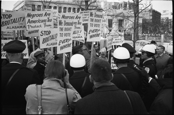 Black-and-white photograph of a dense crowd of protesters holding signs reading "Stop Brutality in Alabama" and "We Demand the Right to Vote Every-Where," with helmeted police officers nearby and a neoclassical building in the background