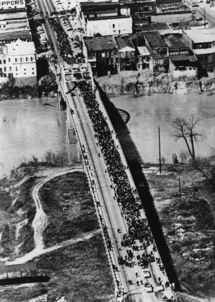 Black-and-white aerial photograph of a dense crowd of marchers crossing a long bridge over a river, with a town visible at the far end