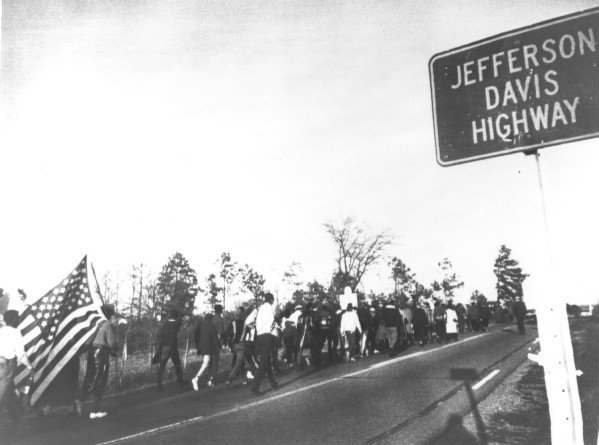 Black-and-white photograph of a large column of marchers walking along a two-lane highway past a road sign reading 'Jefferson Davis Highway.' An American flag is carried at the front of the group