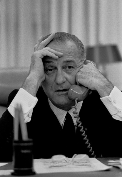 Black-and-white photograph of a man in a dark suit seated at a desk, holding a telephone receiver to his ear with both hands raised to his head