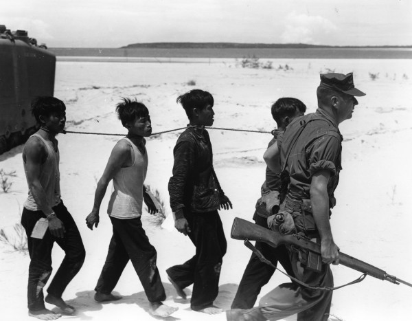 Black-and-white photograph of four barefoot men bound together by a rope around their necks, escorted across a sandy beach by an armed soldier carrying a rifle