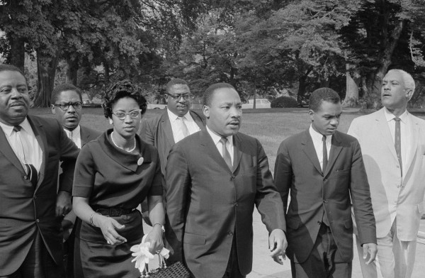 Black-and-white photograph of a group of men and a woman in business attire walking outdoors along a path lined with large trees