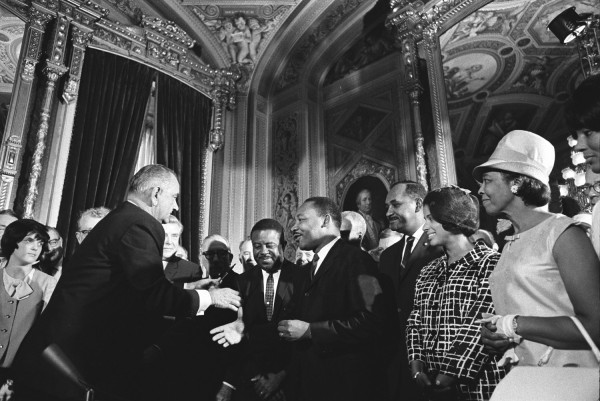 Black-and-white photograph of a man in a dark suit extending a handshake toward a smiling man in a crowd inside an ornately decorated hall