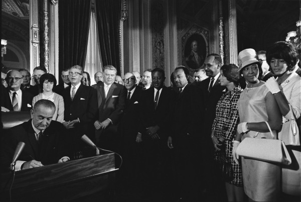 Black-and-white photograph of a man in a suit seated at a desk signing a document while a large crowd of men and women look on in an ornate room