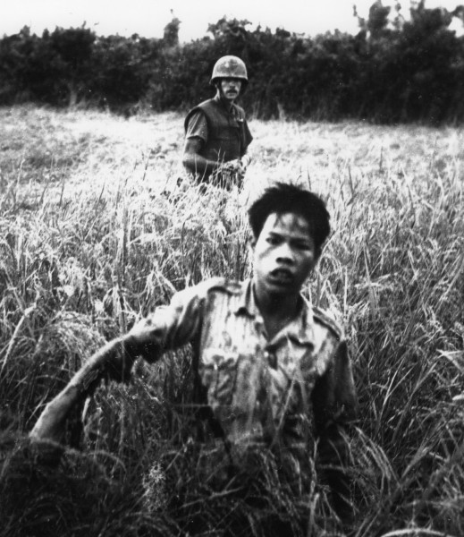 Black-and-white photograph of a man in a light shirt moving through tall grass, with a helmeted soldier standing behind him holding a rope