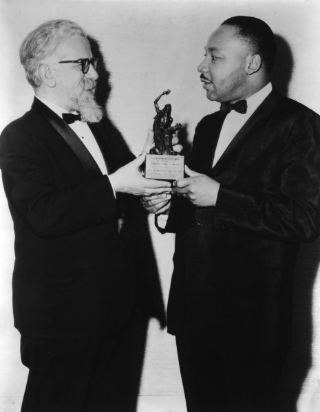 Black-and-white photograph of two men in tuxedos jointly holding a small figurine trophy with an engraved plaque, facing each other