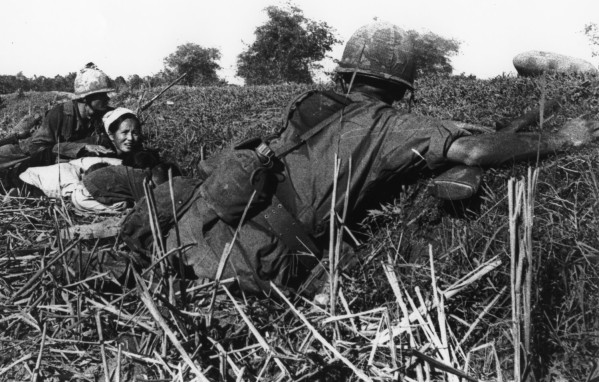 Black-and-white photograph of two helmeted soldiers and a woman lying low in a field of tall grass and dry stalks, trees visible on the horizon