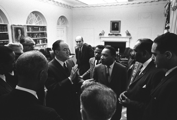 Black-and-white photograph of a group of men and one woman in business attire gathered in a formal room with a fireplace, arched windows, and bookshelves, engaged in conversation