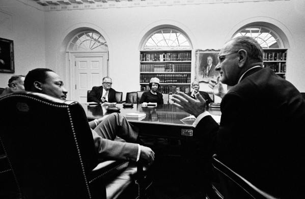 Black-and-white photograph of six people seated around a large conference table in a formal room with arched windows, bookshelves, and a portrait on the wall