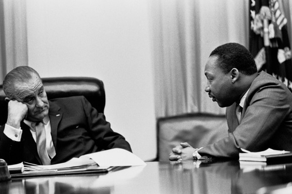 Black-and-white photograph of two men seated across a large conference table, one resting his head on his hand, the other with hands clasped, facing each other