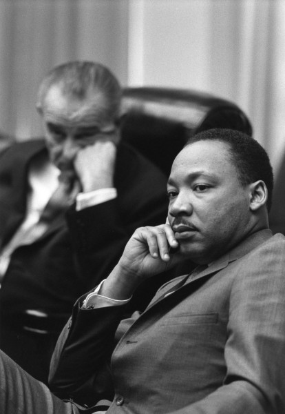 Black-and-white photograph of two men in suits seated in leather chairs; the man in the foreground rests his chin on his hand, looking forward