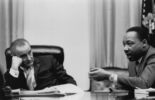 Black-and-white photograph of two men in suits seated across a large table; the man on the left rests his cheek on his fist while the man on the right gestures and speaks
