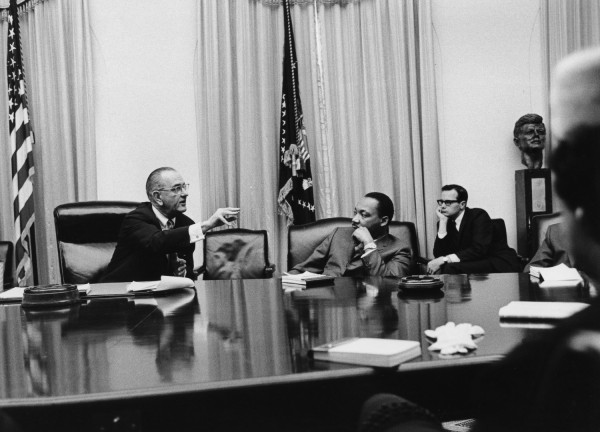 Black-and-white photograph of four men seated around a large conference table; one gestures while speaking, a bust and U.S. flag visible in the background