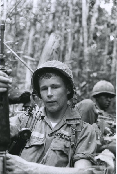 Black-and-white photograph of a young soldier in a helmet and U.S. Army uniform in a jungle setting, with a second helmeted soldier visible behind him
