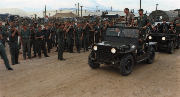 Color photograph of two military jeeps moving past a large crowd of soldiers in green fatigues at a dusty outdoor base, with Quonset huts and mountains visible in the background