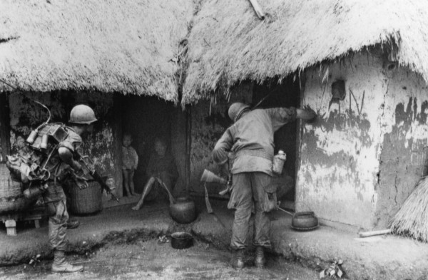 Black-and-white photograph of two helmeted soldiers in full gear approaching a thatched-roof hut; two children are visible sheltering inside the doorway