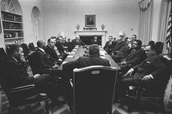 Black-and-white photograph of a large group of men in suits seated around a long conference table in a formal room with a fireplace and portrait above it