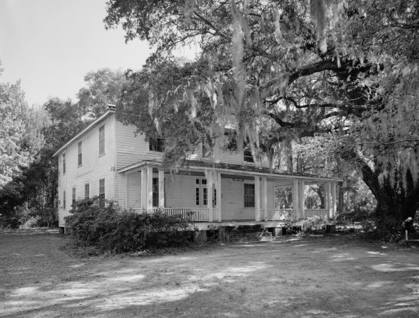 Black-and-white photograph of a two-story white clapboard building with a wide covered porch, surrounded by large oak trees draped with Spanish moss