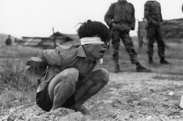Black-and-white photograph of a blindfolded man with hands bound behind his back, crouching on bare ground and crying out, with uniformed soldiers standing in the background