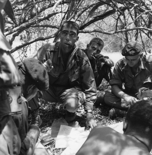 Black-and-white photograph of several soldiers in camouflage uniforms crouching under tree branches, gathered around a map on the ground