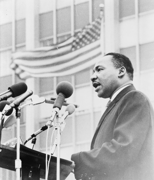 Black-and-white photograph of a man in a suit speaking at an outdoor podium lined with multiple microphones, an American flag visible behind him