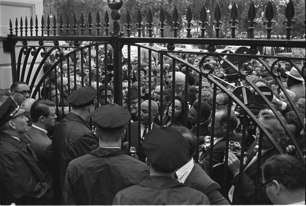 Black-and-white photograph of a large crowd pressing against ornate iron gates, with police officers on one side and protesters and camera operators on the other