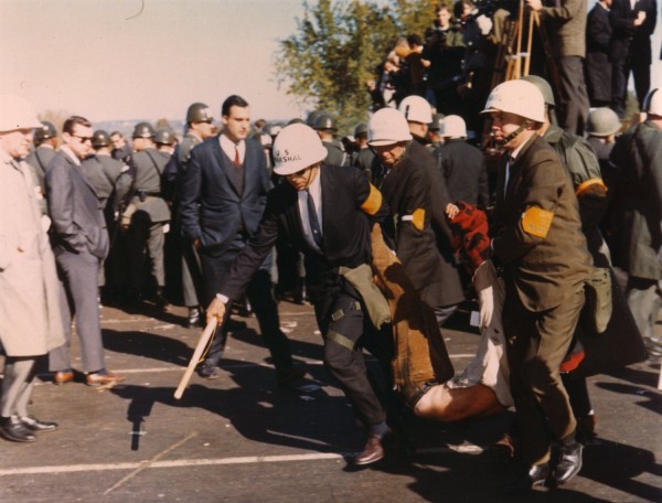 Color photograph of U.S. Marshals in white hard hats dragging a limp protester across pavement while soldiers and onlookers crowd the background