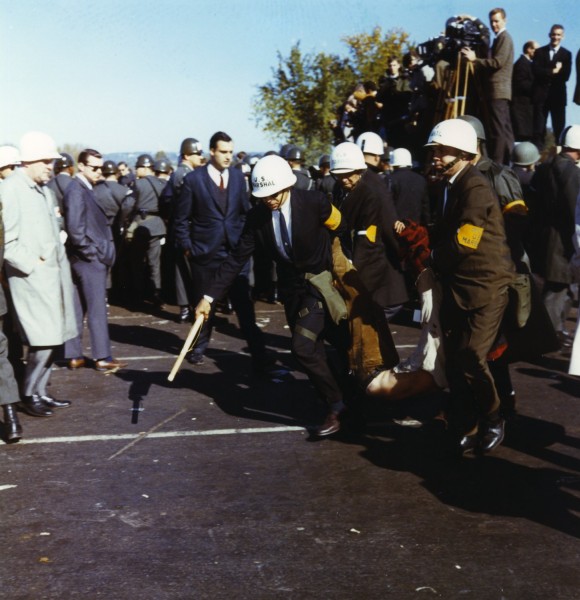 Color photograph of U.S. Marshals in white helmets dragging a limp demonstrator across pavement while a crowd and news photographers look on