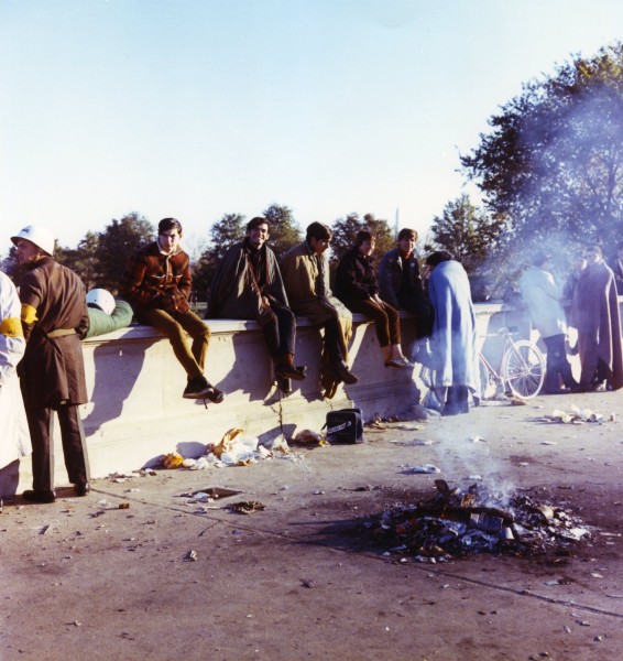 Color photograph of several young people seated on a low concrete wall outdoors, a small fire burning on the pavement in the foreground, trees and a monument visible in the background