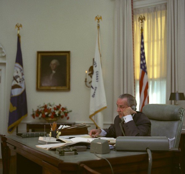 Color photograph of a man in a dark suit seated at a large desk, holding a telephone receiver, with flags and a framed portrait visible behind him