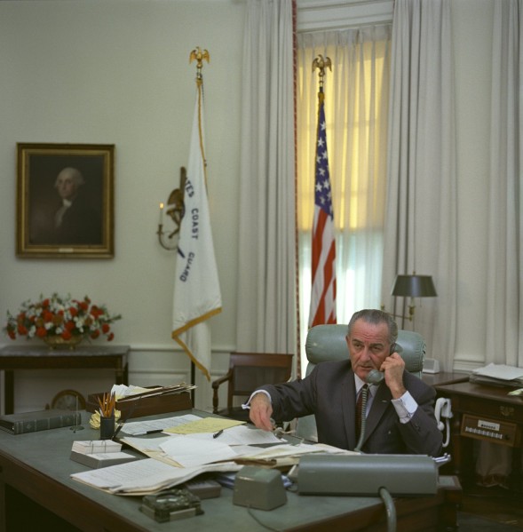 Color photograph of a man in a suit seated at a large desk covered in papers, holding a telephone receiver, with a U.S. flag and Coast Guard flag behind him