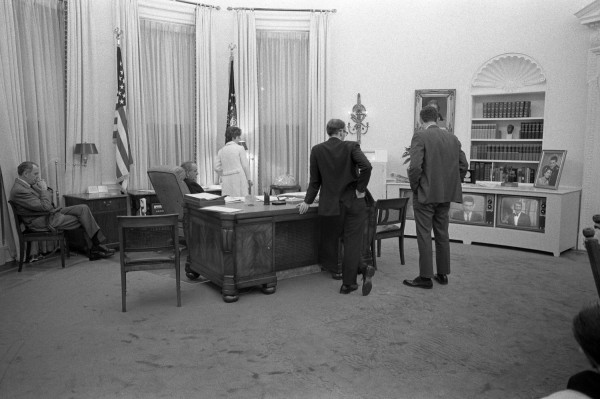Black-and-white photograph of several people gathered in a large oval-shaped office around a large desk, watching a television set displaying a news broadcast in the background