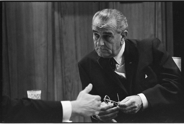 Black-and-white photograph of an older man in a dark suit holding eyeglasses, seated at a table, with a glass visible at left and a second person's hand extended toward him