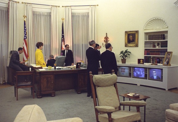 Color photograph of a group of men and a woman in a yellow coat gathered around a large desk in a formal office, facing three television sets displaying a 'Special Report' broadcast