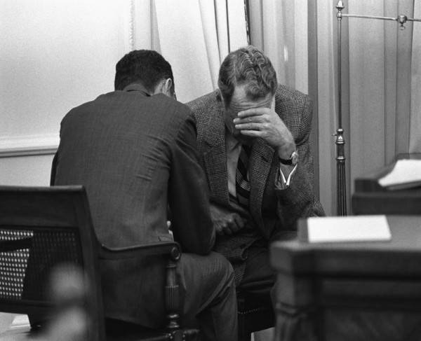 Black-and-white photograph of two men in suits seated close together in an office; one buries his face in his hand while the other leans toward him